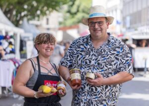 Deux producteurs du Confit d'Ascain au Pays Basque tenant des produits artisanaux, tels que des confitures et conserves, dans une ambiance de marché convivial.